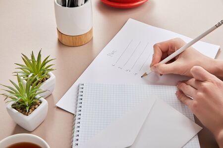 cropped view of woman writing plan on paper near green plants, cup of tea, envelope, blank notebook...