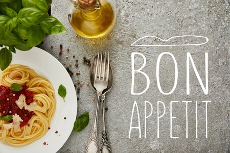 top view of delicious spaghetti with tomato sauce on plate near basil leaves, oil and cutlery on grey textured surface with bon appetit illustrationの写真素材