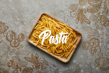 top view of raw penne on wooden board on grey textured surface with pasta illustrationの写真素材