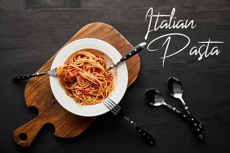 top view of tasty bolognese pasta with tomato sauce in white plate on wooden cutting board near cutlery on black wooden background, italian pasta illustrationの写真素材