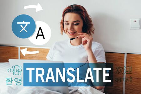 smiling girl making video call with headset and laptop in bed during quarantine with translate letteringの写真素材