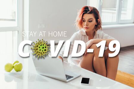 beautiful woman with headphones using laptop on kitchen with apples and smartphone during quarantine with stay at home COVID-19 letteringの写真素材