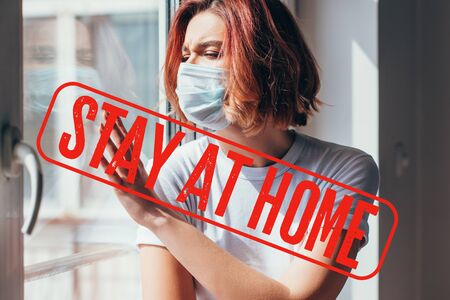 upset girl in medical mask looking through window during self isolation with stay at home letteringの写真素材