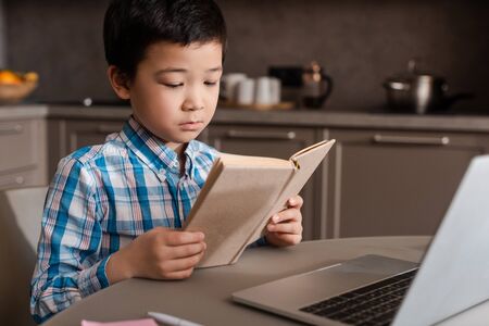 asian boy reading book and studying online with laptop at home during self isolation