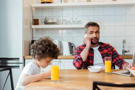selective focus of freelancer father talking on smartphone and looking at curly son in kitchenの写真素材