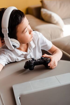 sad asian boy playing video game with joystick and laptop on self isolation