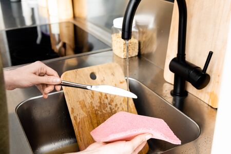Cropped view of woman holding rag and knife near kitchen faucet and sinkの写真素材