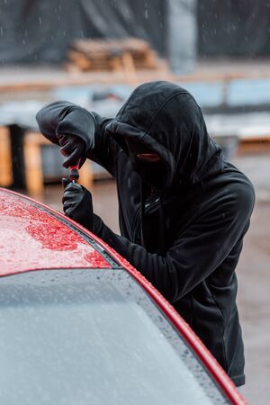 Selective focus of burglar in mask opening door of car with screwdriver during rain on urban streetの写真素材