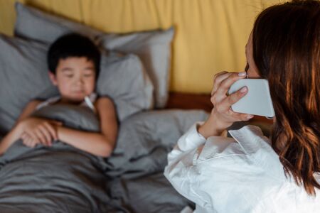 diseased asian boy lying in bed while mother calling to doctor on quarantineの写真素材