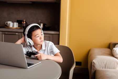 KYIV, UKRAINE - APRIL 22, 2020: bored asian boy playing video game with joystick and laptop on...