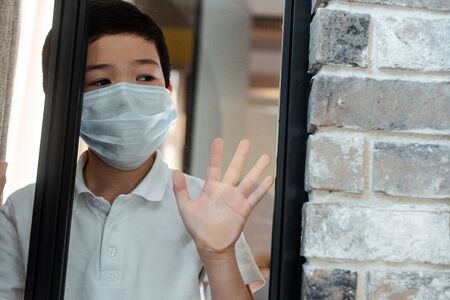 sad asian boy in medical mask looking through window on quarantineの写真素材