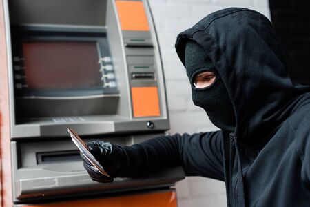 Side view of robber in balaclava holding credit cards near automated teller machine