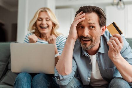 Confused man holding credit card near cheerful wife using laptop at home