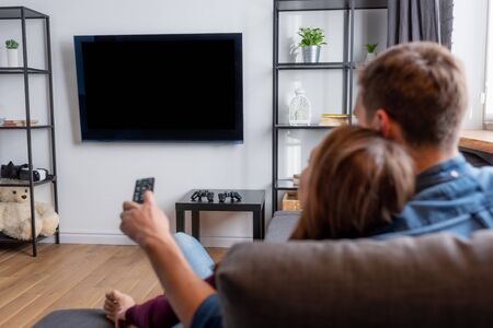 selective focus of man holding remote controller and looking at flat panel tv with blank screen near woman in living roomの写真素材