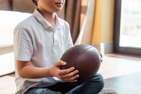 cropped view of asian boy with rugby ball sitting on floor at home on quarantineの写真素材