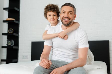 curly son hugging cheerful and bearded father in bedroom