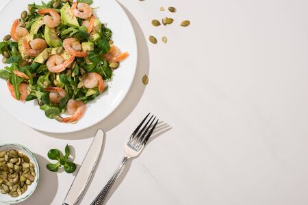 top view of fresh green salad with pumpkin seeds, shrimps and avocado on plate near cutlery on white backgroundの写真素材