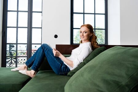 Side view of woman smiling at camera while using digital tablet on couch