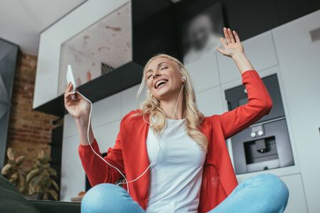 low angle view of happy woman in earphones singing with closed eyes while holding smartphone