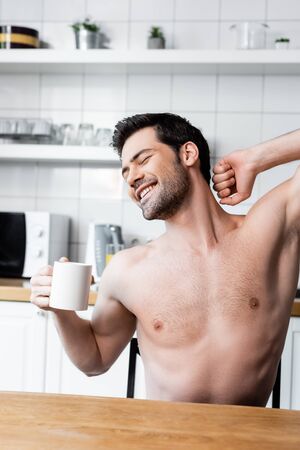 happy shirtless man stretching and holding cup of coffee on kitchen in morningの写真素材