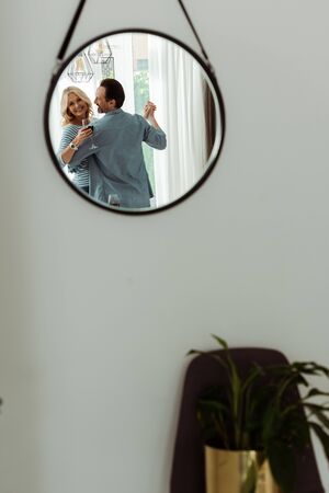 Selective focus of smiling woman reflecting in mirror while holding glass of wine and dancing with husband at homeの写真素材