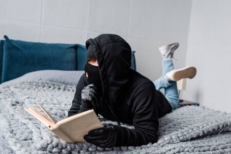 Selective focus of thief in balaclava and leather gloves reading book on bedの写真素材