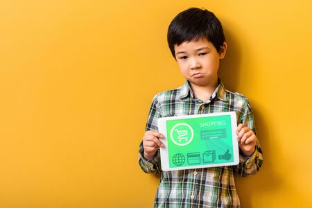 upset asian boy holding digital tablet with shopping app on yellowの写真素材