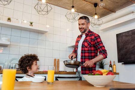 selective focus of happy boy looking at cheerful father holding frying pan in kitchenの写真素材