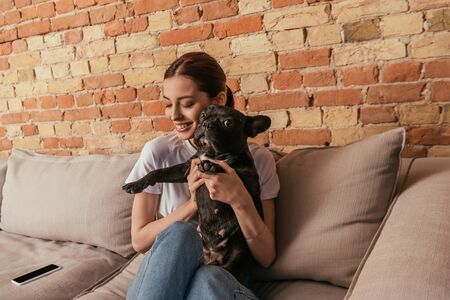 happy girl holding in arms cute french bulldog while sitting on sofa near smartphone with blank screenの写真素材