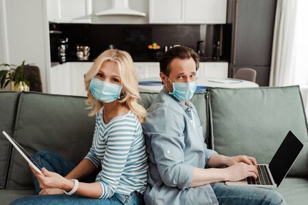 Side view of mature couple in medical masks looking at camera while using gadgets on couchの写真素材