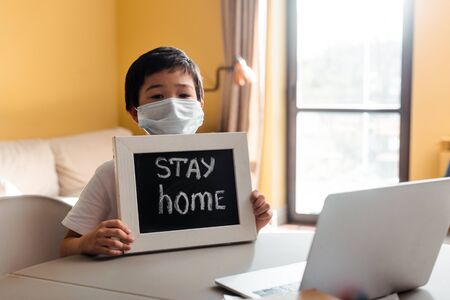 asian boy in medical mask holding board with stay home lettering in front of laptop during self isolationの写真素材