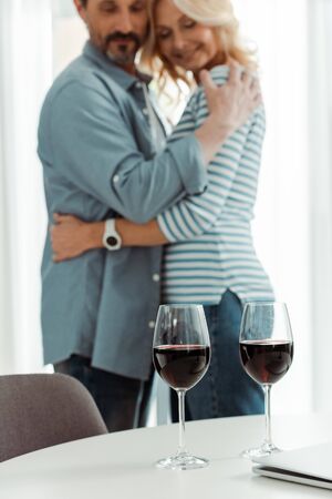Selective focus of glasses of wine and laptop on table near smiling mature couple embracing in kitchenの写真素材