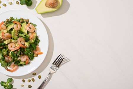 top view of fresh green salad with pumpkin seeds, shrimps and avocado on plate near fork and ingredients on white backgroundの写真素材