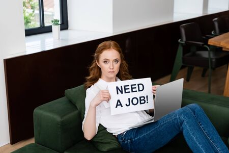 Attractive freelancer looking at camera while holding card with need a job lettering and laptop on...