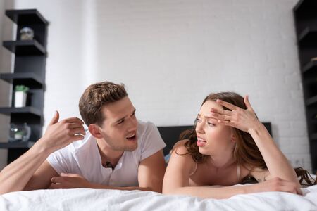 displeased couple looking at each other while lying on bed and feeling hotの写真素材