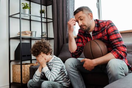 displeased man holding basketball while watching championship with upset son covering faceの写真素材