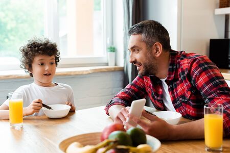 selective focus of happy father holding smartphone near surprised son in kitchen