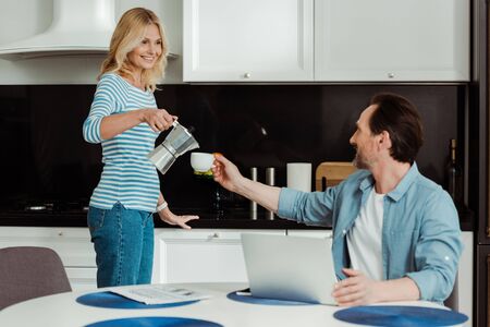 Selective focus of smiling woman pouring coffee near husband and laptop on kitchen tableの写真素材