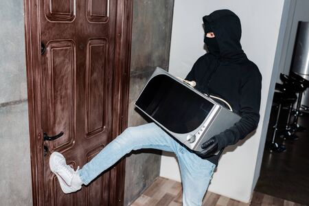 Thief in mask holding microwave and closing house door during stealingの写真素材