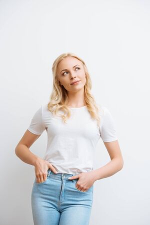dreamy, smiling woman looking away while holding hands in pockets isolated on whiteの写真素材
