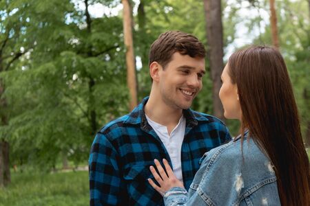cheerful student looking at girlfriend in denim jacket in park