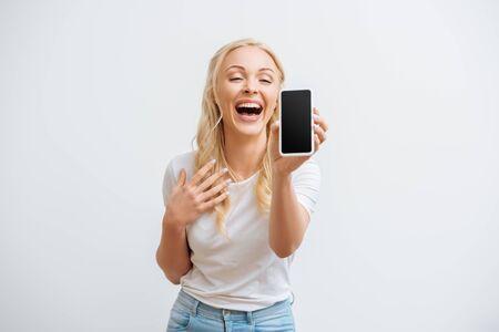 excited girl laughing while showing smartphone with blank screen isolated on whiteの写真素材