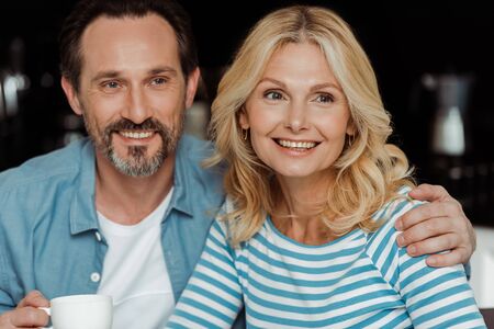 Handsome man holding cup of coffee and embracing smiling wife at home
