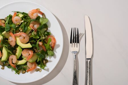 top view of fresh green salad with pumpkin seeds, shrimps and avocado on plate near cutlery on white backgroundの写真素材