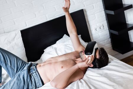 shirtless man gesturing while lying and using virtual reality headset on bedの写真素材
