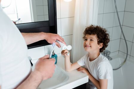 selective focus of father holding toothpaste near toothbrush of cheerful and curly sonの写真素材