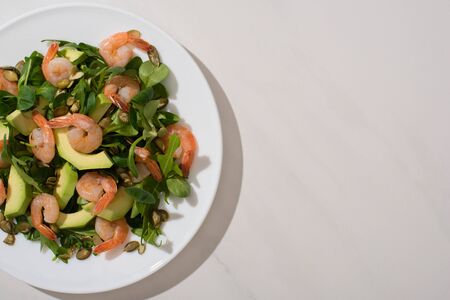 top view of fresh green salad with pumpkin seeds, shrimps and avocado on plate on white backgroundの写真素材