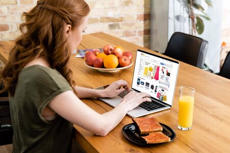 KYIV, UKRAINE - MAY 8, 2020 : Side view of woman using laptop with ebay website near glass of orange juice and toasts with jam on tableの写真素材