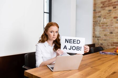 Woman looking at camera while holding card with need a job lettering near laptop on table
