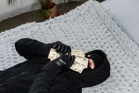 Robber in balaclava holding dollars near face while lying on bed